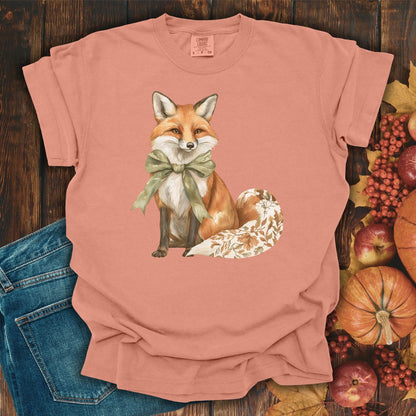 Terracotta garment-dyed cotton t-shirt with a watercolor fox wearing a sage green bow and leaf-patterned tail.