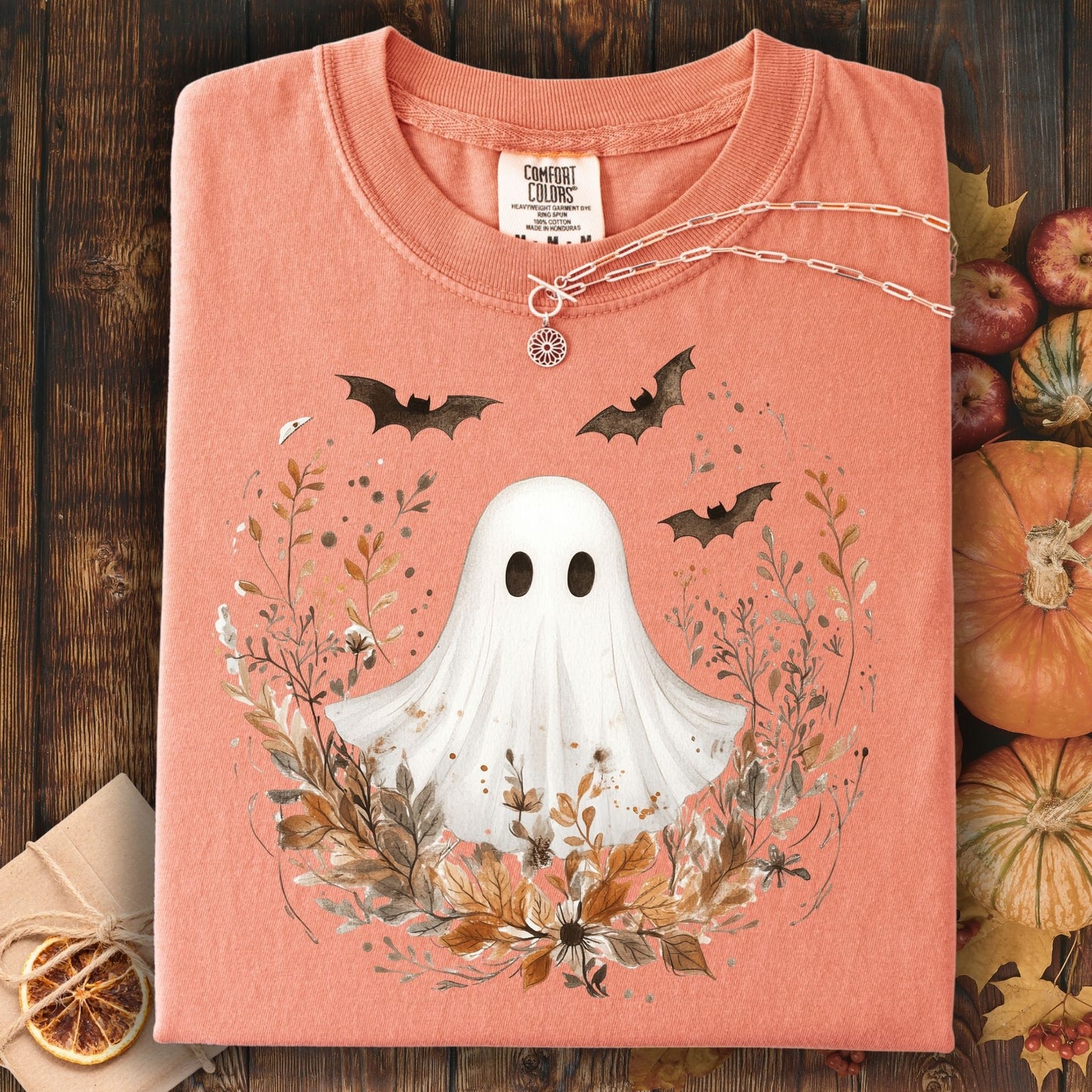 Folded light orange t-shirt with ghost and leaf design on a wooden surface with pumpkins.