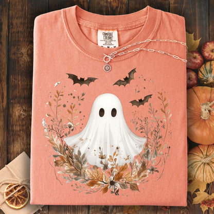 Folded light orange t-shirt with ghost and leaf design on a wooden surface with pumpkins.
