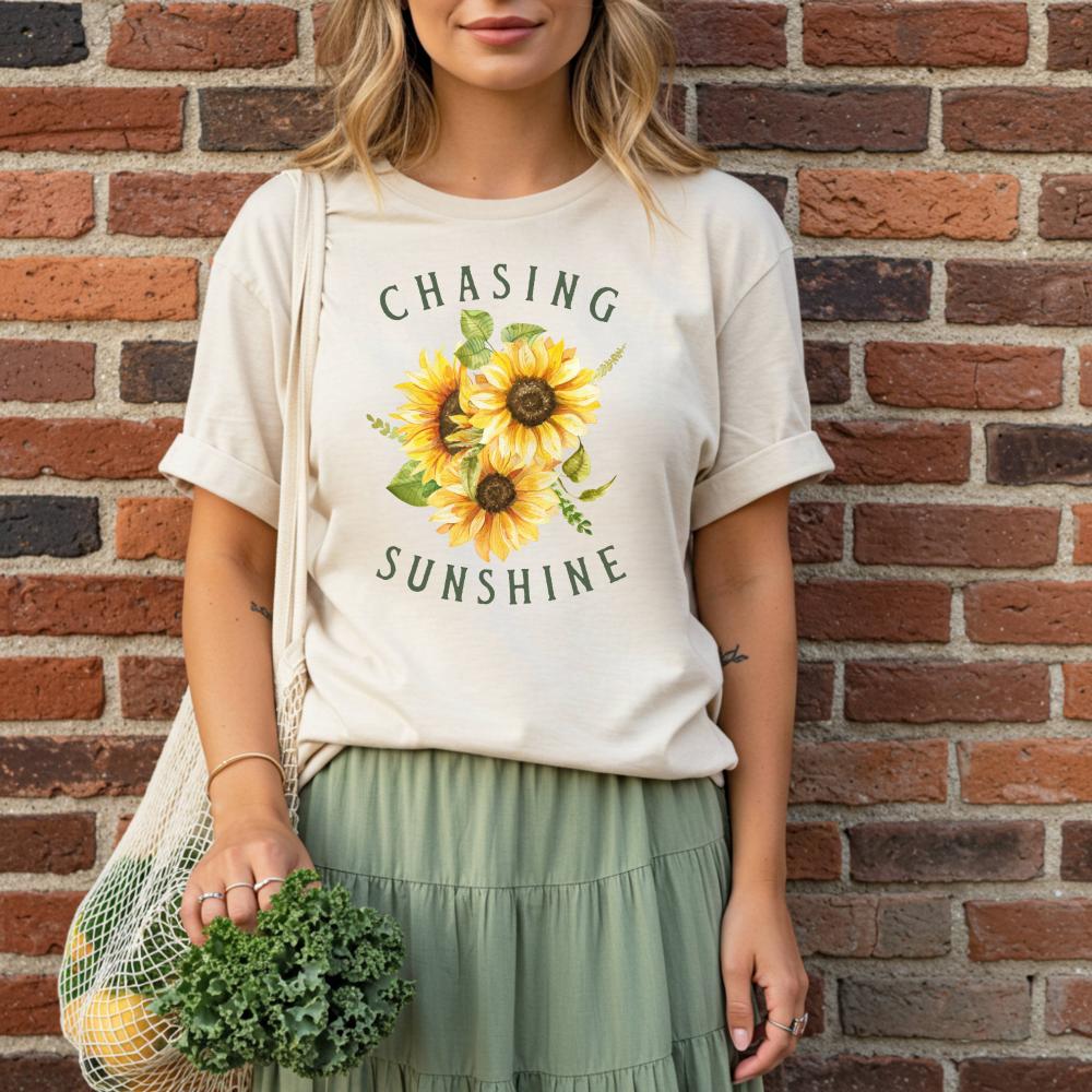 woman wearing a t-shirt with sunflower design and text 'Chasing Sunshine' against a brick wall.