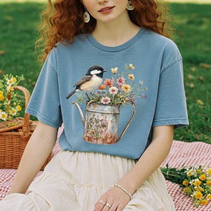 Garment-dyed blue t-shirt with a chickadee perched on a floral watering can, worn by a person on a picnic blanket.