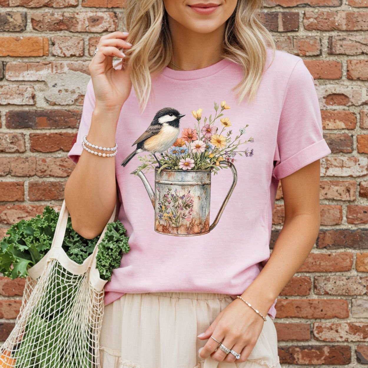 Pink garment-dyed t-shirt with a chickadee perched on a floral watering can, worn by a woman holding kale.