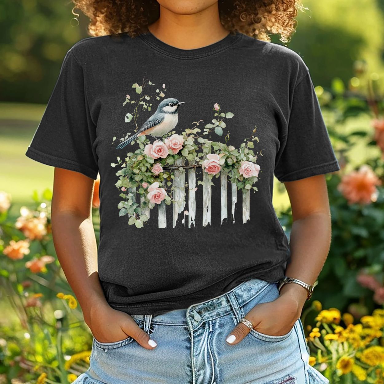 Garment-dyed black t-shirt with a watercolor chickadee on a weathered white picket fence with pink roses.
