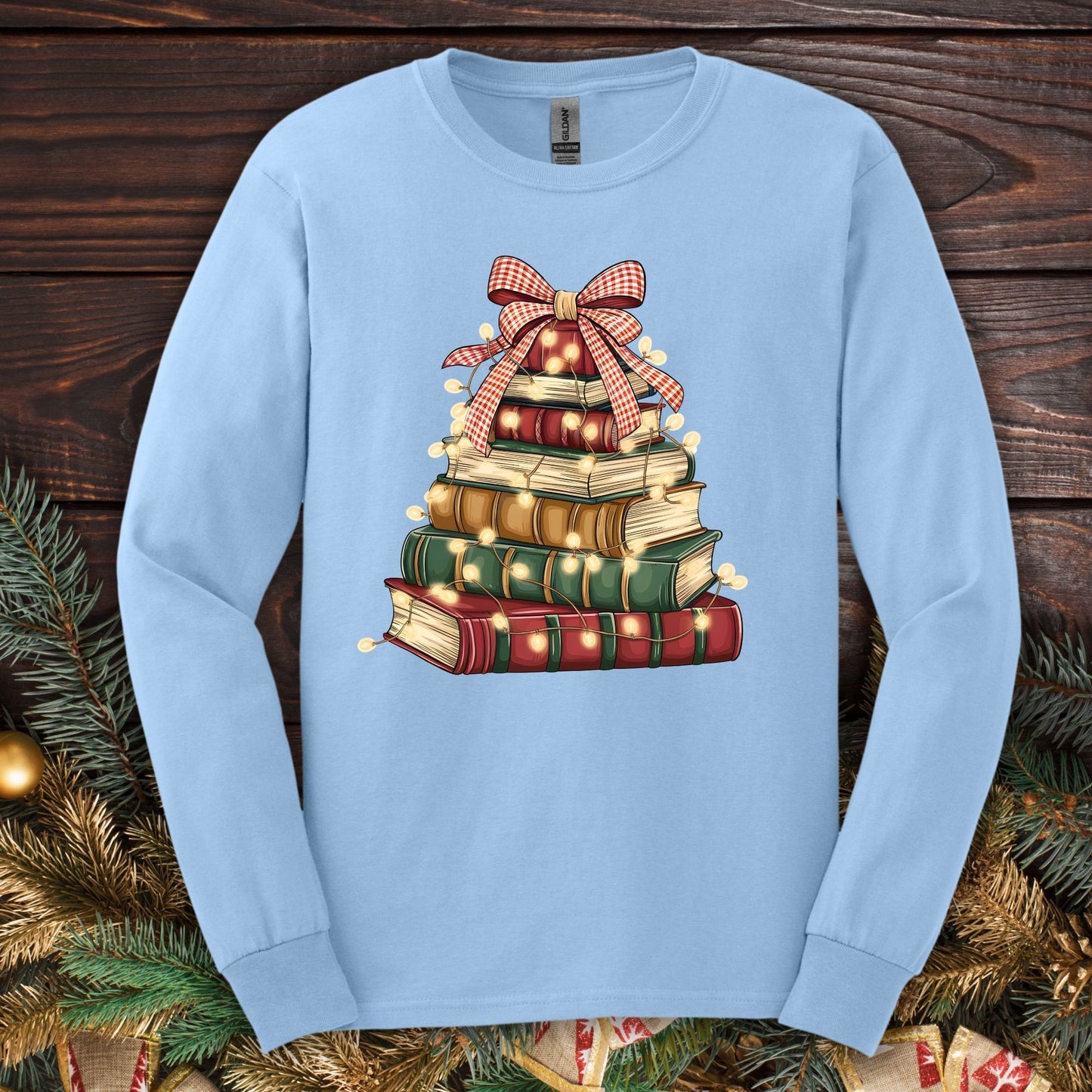 Light blue long sleeve tee with a watercolor Christmas book tree design, topped with a plaid bow and string lights.
