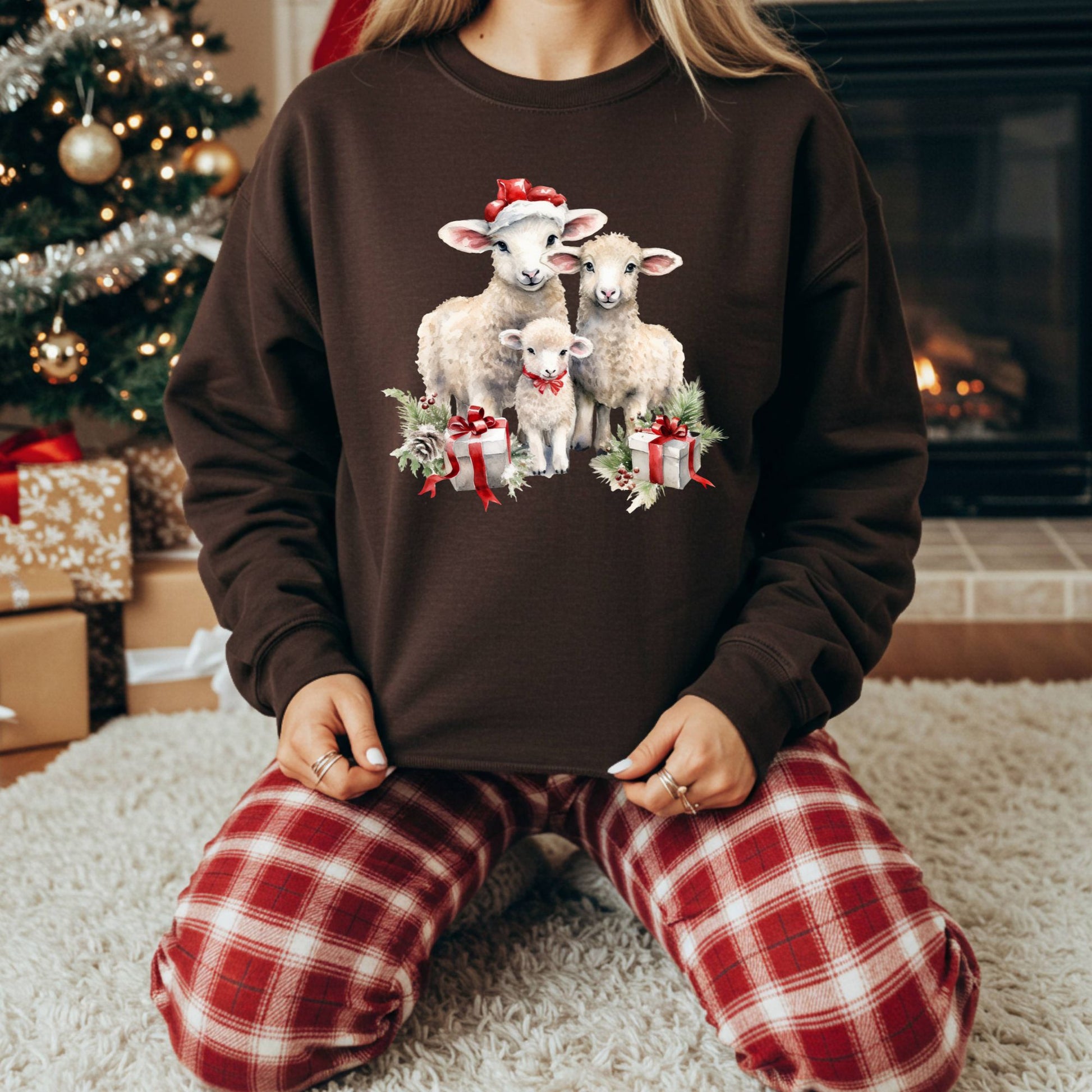 Brown crewneck sweatshirt with three watercolor lambs in Santa hats and gifts, worn by a person in plaid pajamas.