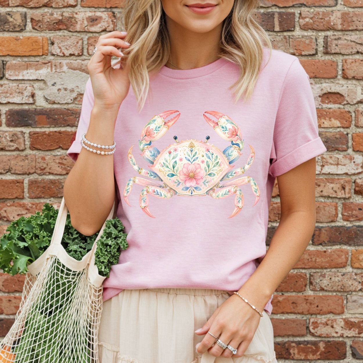 Pink garment-dyed t-shirt with a watercolor floral crab design, worn by a woman holding a mesh bag of greens.