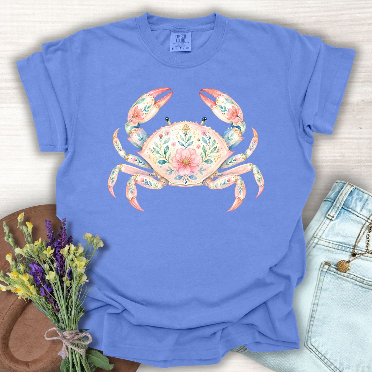 Periwinkle S t-shirt with a watercolor floral crab design, next to jeans and flowers.