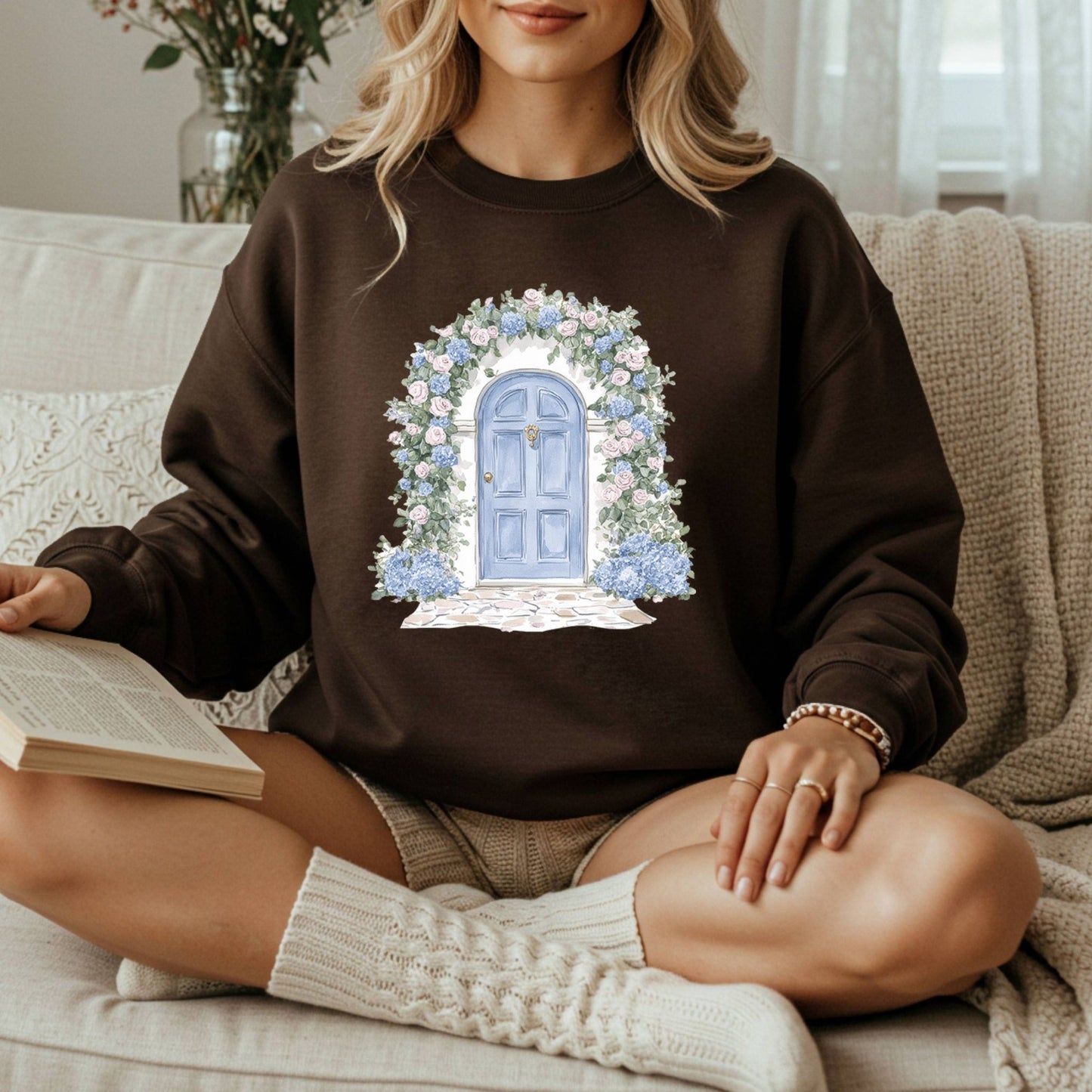 Woman in brown sweatshirt with watercolor cottage doorway, pink roses, and blue hydrangeas.