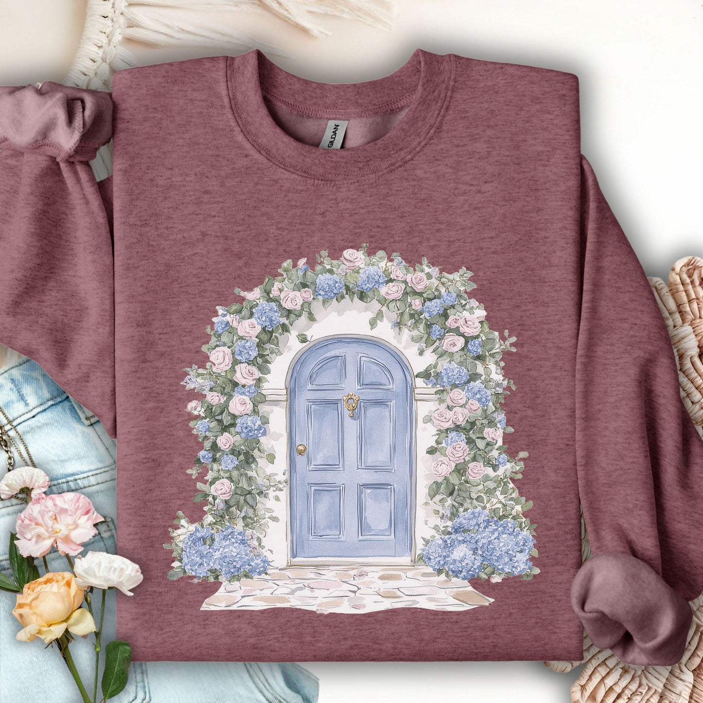 Heather Sport Dark Maroon sweatshirt with a watercolor cottage doorway framed by blue hydrangeas and pink roses.