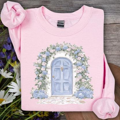 Light pink sweatshirt with a watercolor cottage doorway, blue door, and pink/blue hydrangeas.