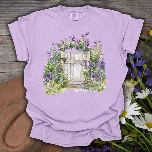 Orchid S-size garment-dyed t-shirt with a watercolor garden gate and purple wildflowers.