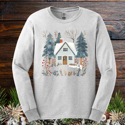 Ash long sleeve tee with a hand-drawn winter cabin, pines, and red berries, on a wood background.