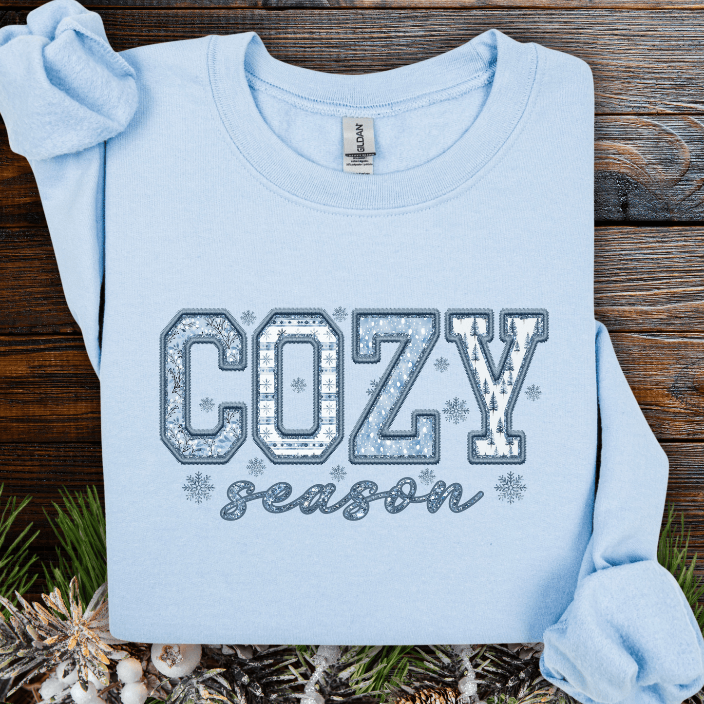 Light blue sweatshirt with "COZY season" faux patchwork design in blue and white, featuring snowflakes and winter patterns.