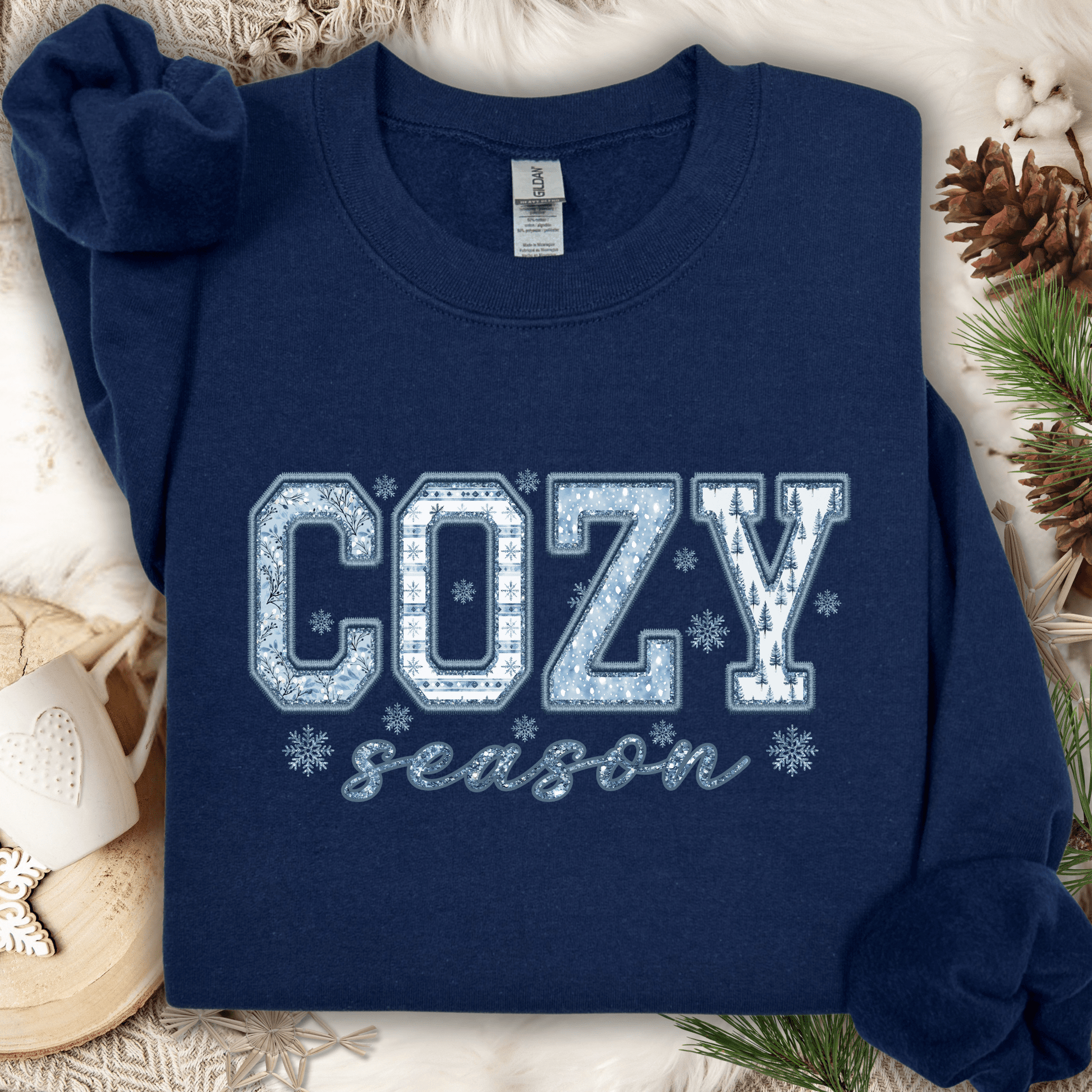 Navy S sweatshirt with "COZY season" faux patchwork print in blue and white, on a cozy winter background.