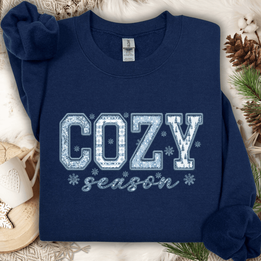 Navy S sweatshirt with "COZY season" faux patchwork print in blue and white, on a cozy winter background.