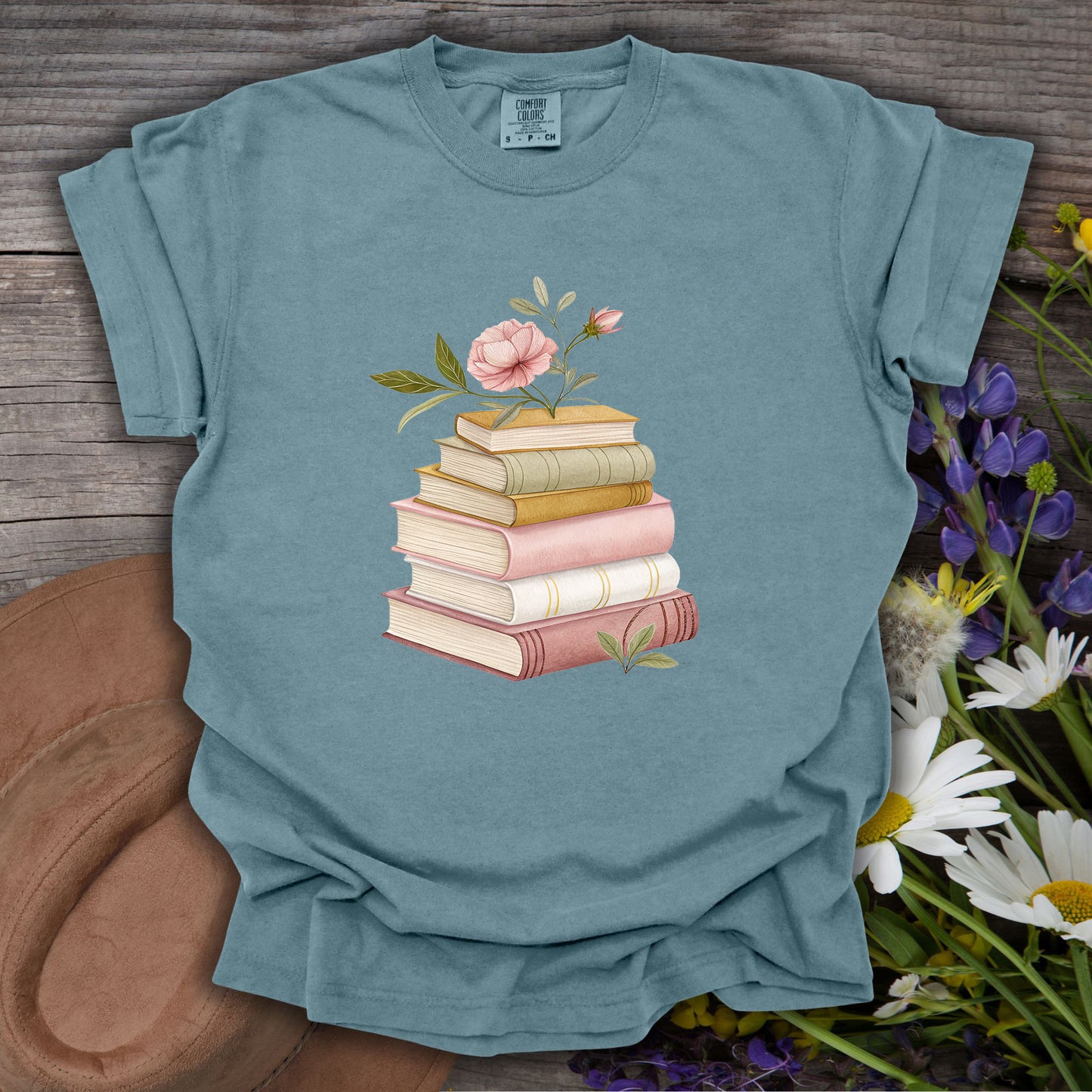 Ice Blue S t-shirt with a watercolor stack of books and pink floral illustration on a wooden background.