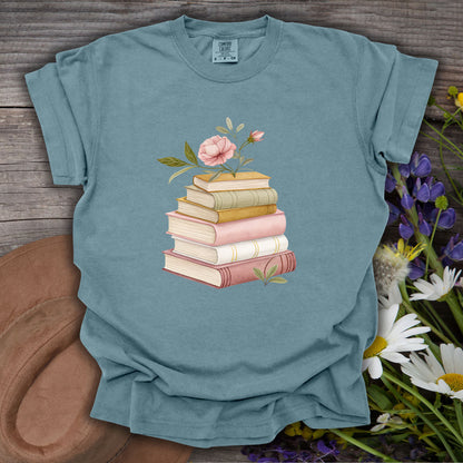 Ice Blue S t-shirt with a watercolor stack of books and pink floral illustration on a wooden background.