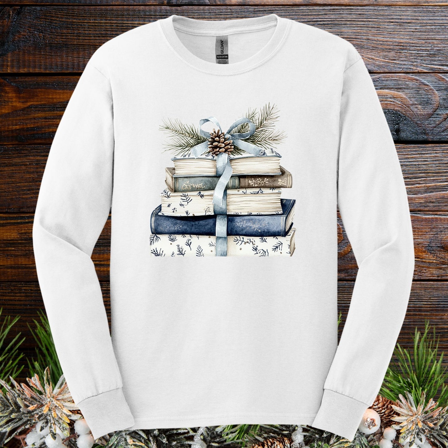 White long-sleeve tee with a stack of blue and white books, ribbon, evergreen, and pinecone design.
