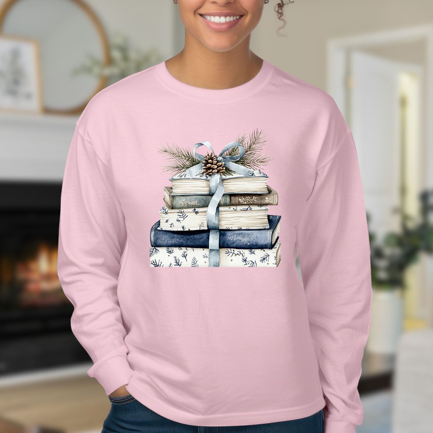 Pink long-sleeve tee with a stack of blue and white books, ribbon, evergreen, and pinecone design.