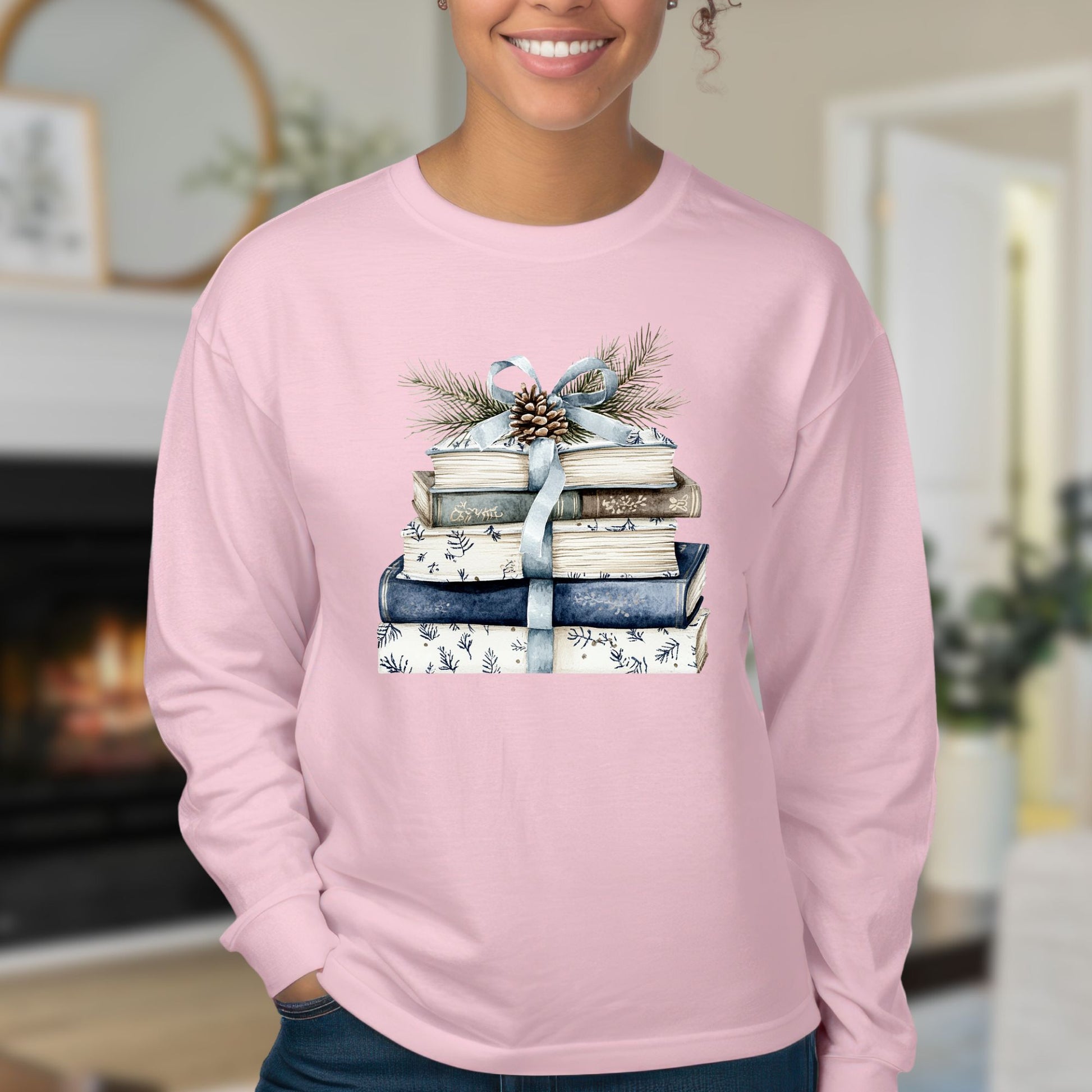 Pink long-sleeve tee with a stack of blue and white books, ribbon, evergreen, and pinecone design.