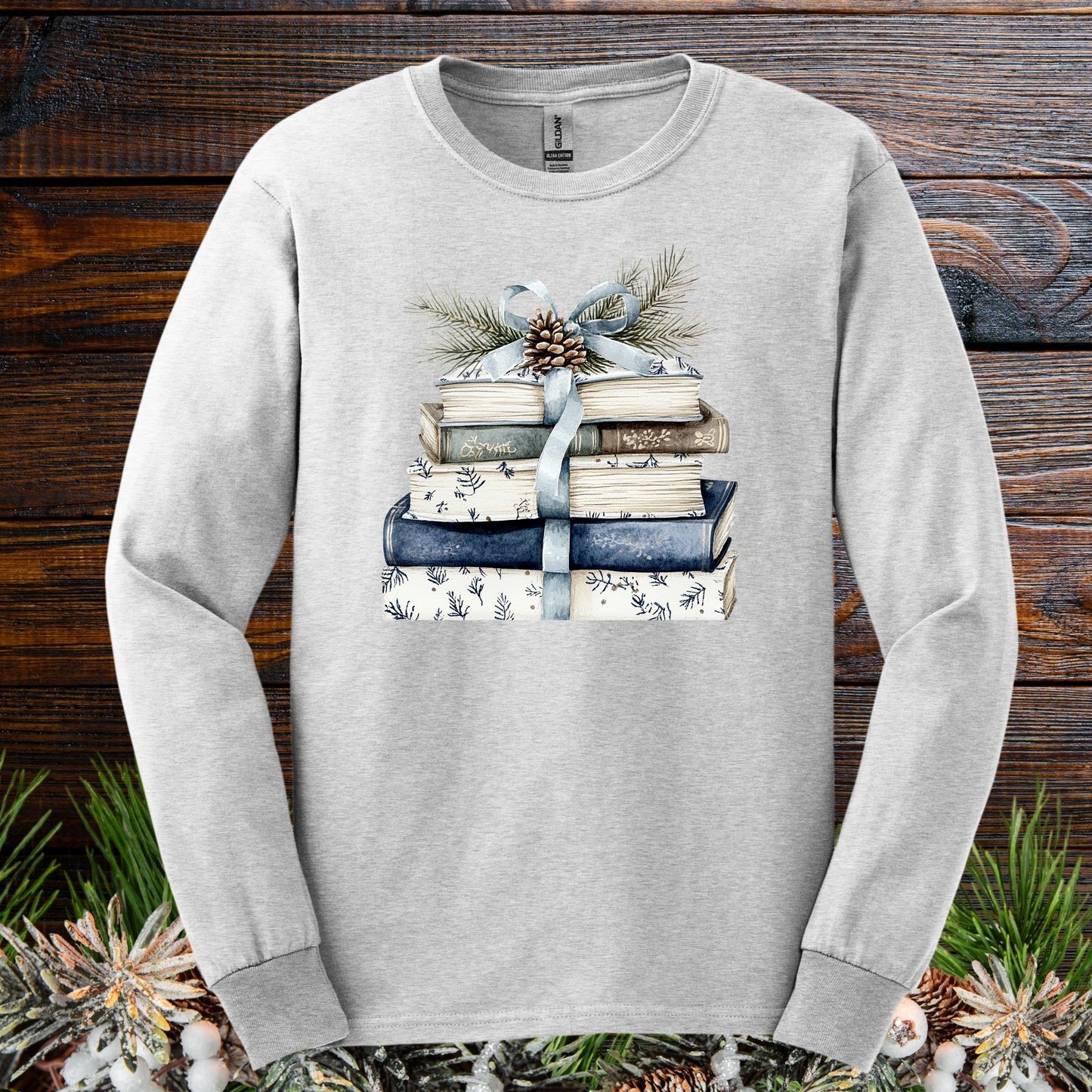 Ash long sleeve tee with a stack of blue and white books, blue ribbon, evergreen, and pinecone design.