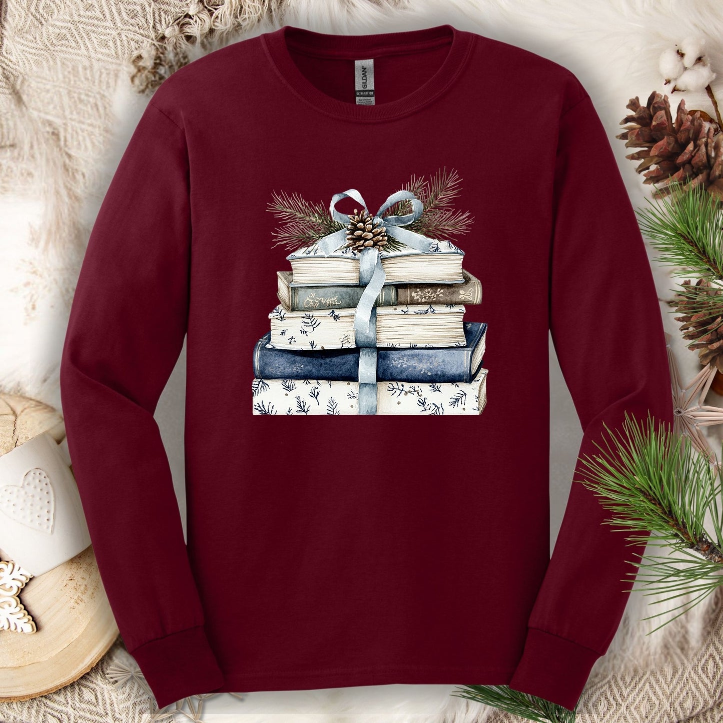 Maroon long-sleeve tee with a stack of blue and white books, blue ribbon, evergreen, and pinecone.