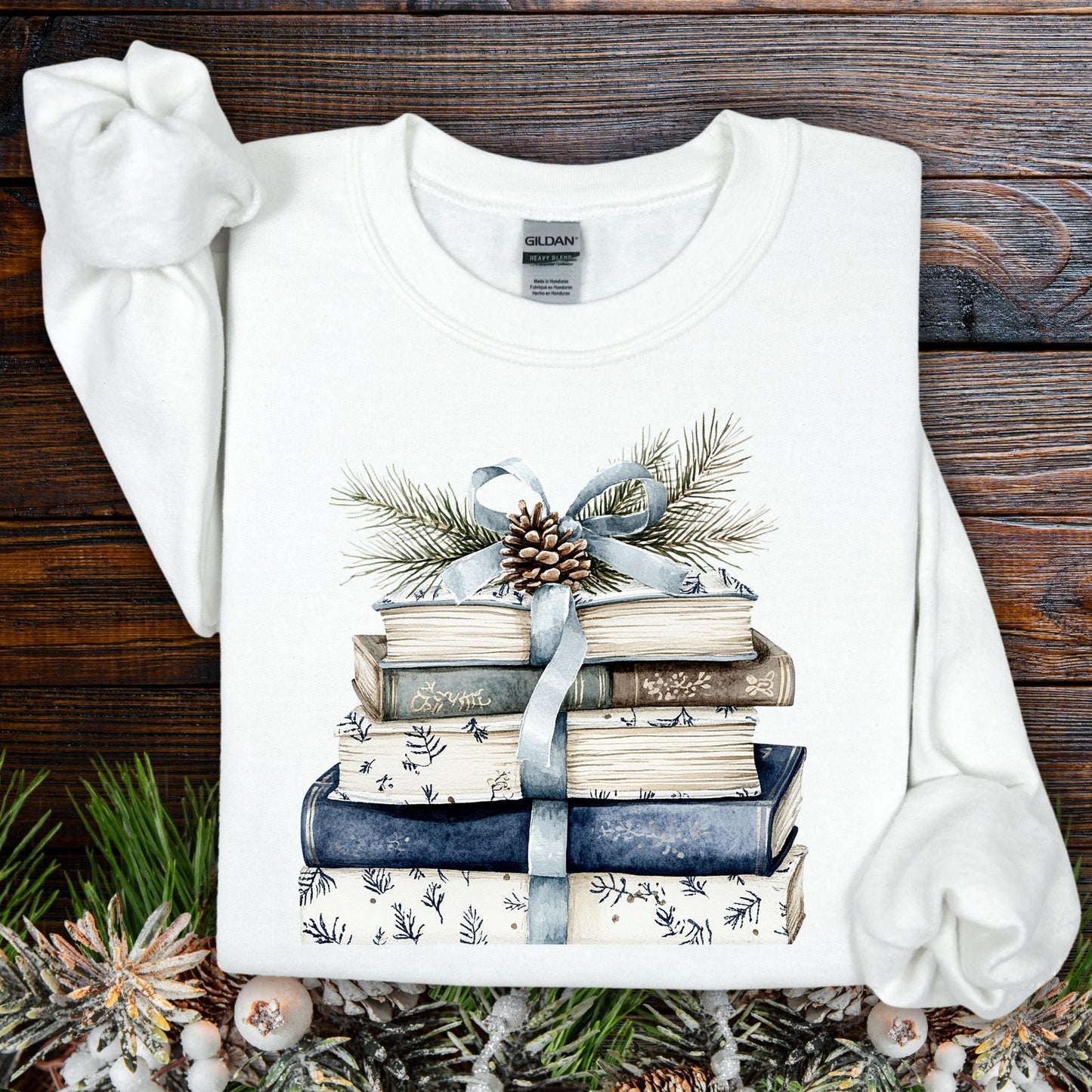 White sweatshirt with a stack of blue and white books, blue ribbon, evergreen, and pinecone design.