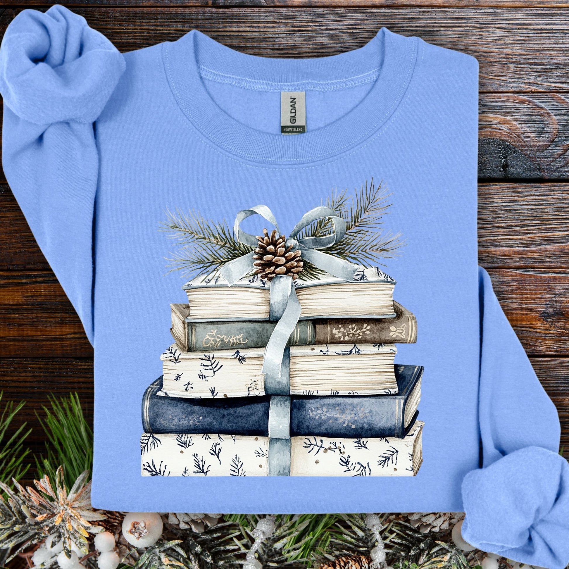 Light blue sweatshirt with a stack of blue and white books, ribbon, evergreen, and pinecone design.