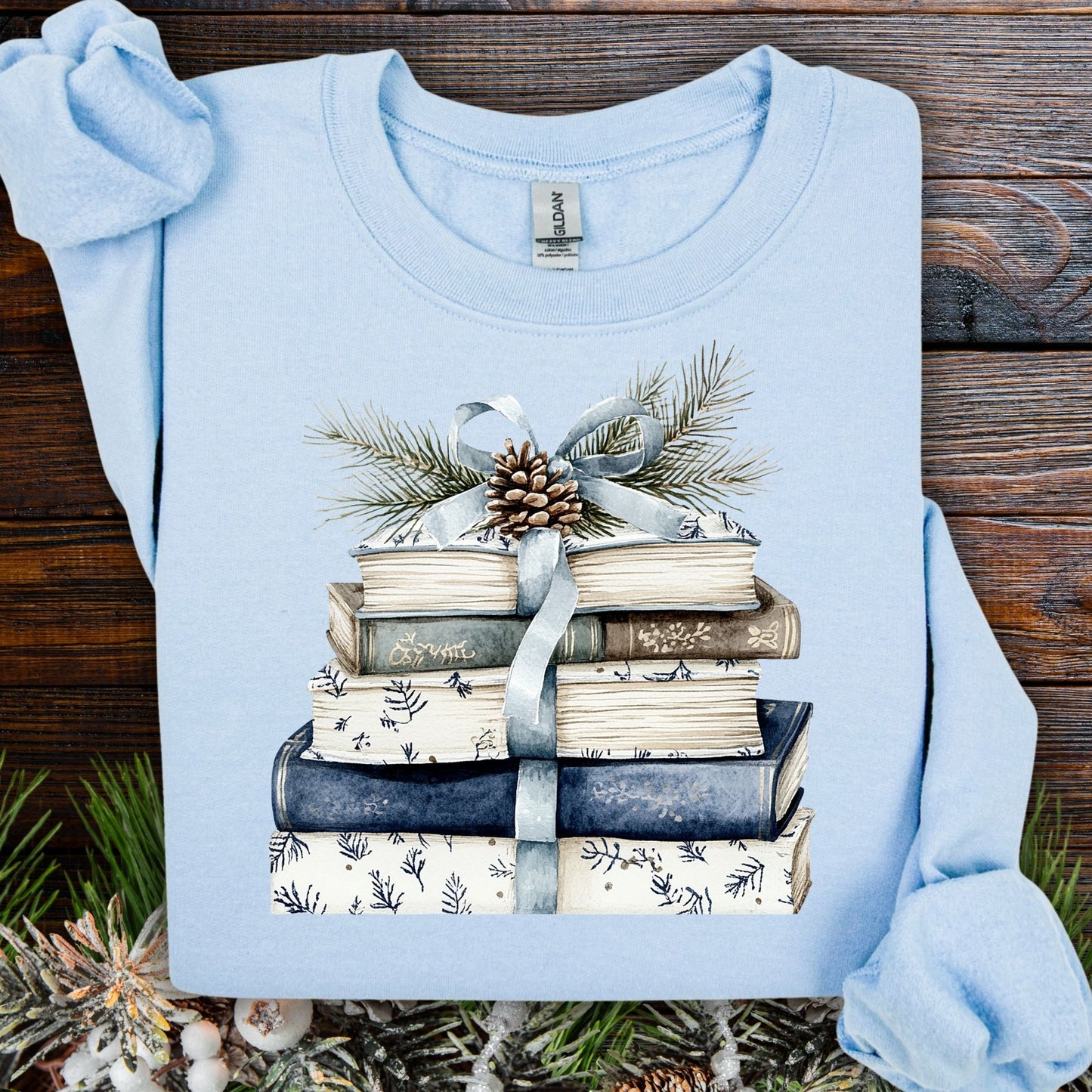 Light blue sweatshirt with a stack of blue and white books, ribbon, evergreen, and pinecone design.
