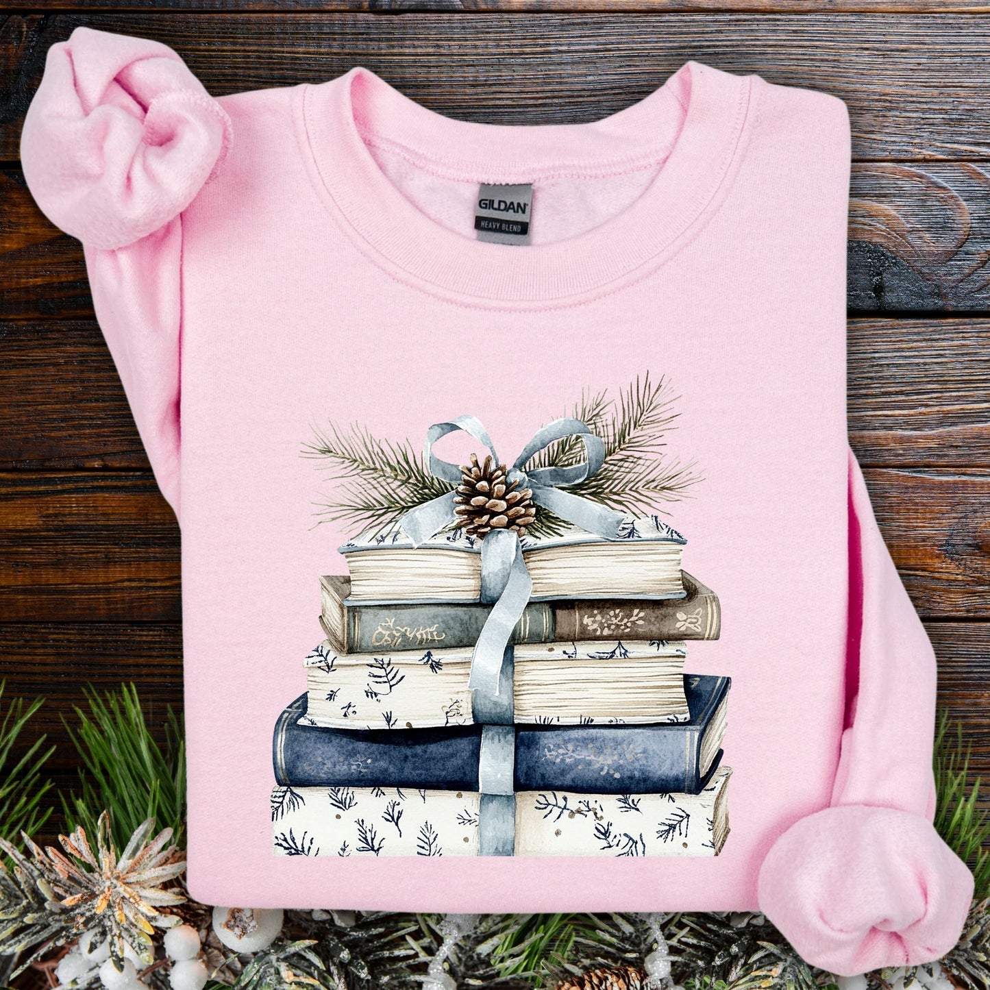 Light pink Gildan sweatshirt with a stack of blue and white books, ribbon, evergreen, and pinecone design.