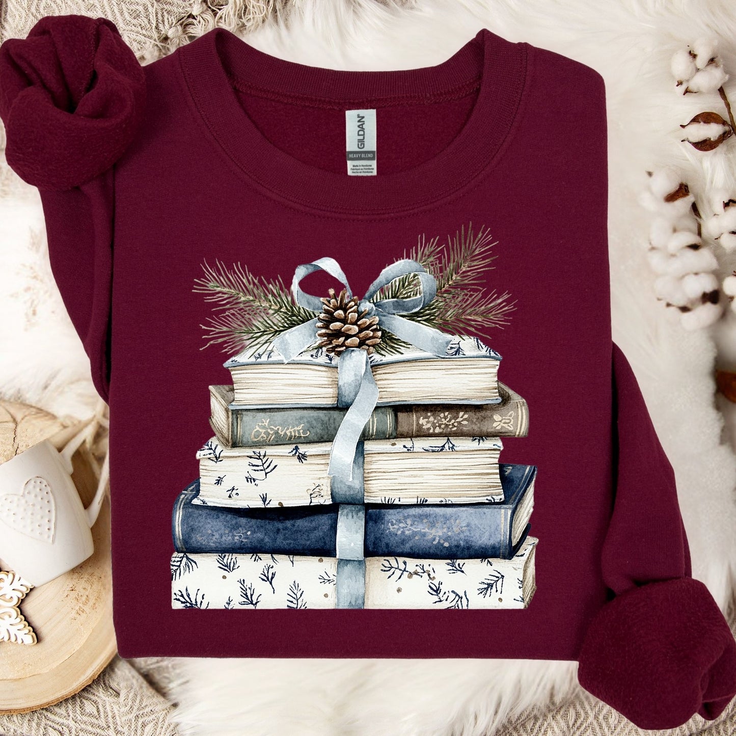 Maroon Gildan sweatshirt with a stack of blue and white books, blue ribbon, evergreen, and pinecone design.
