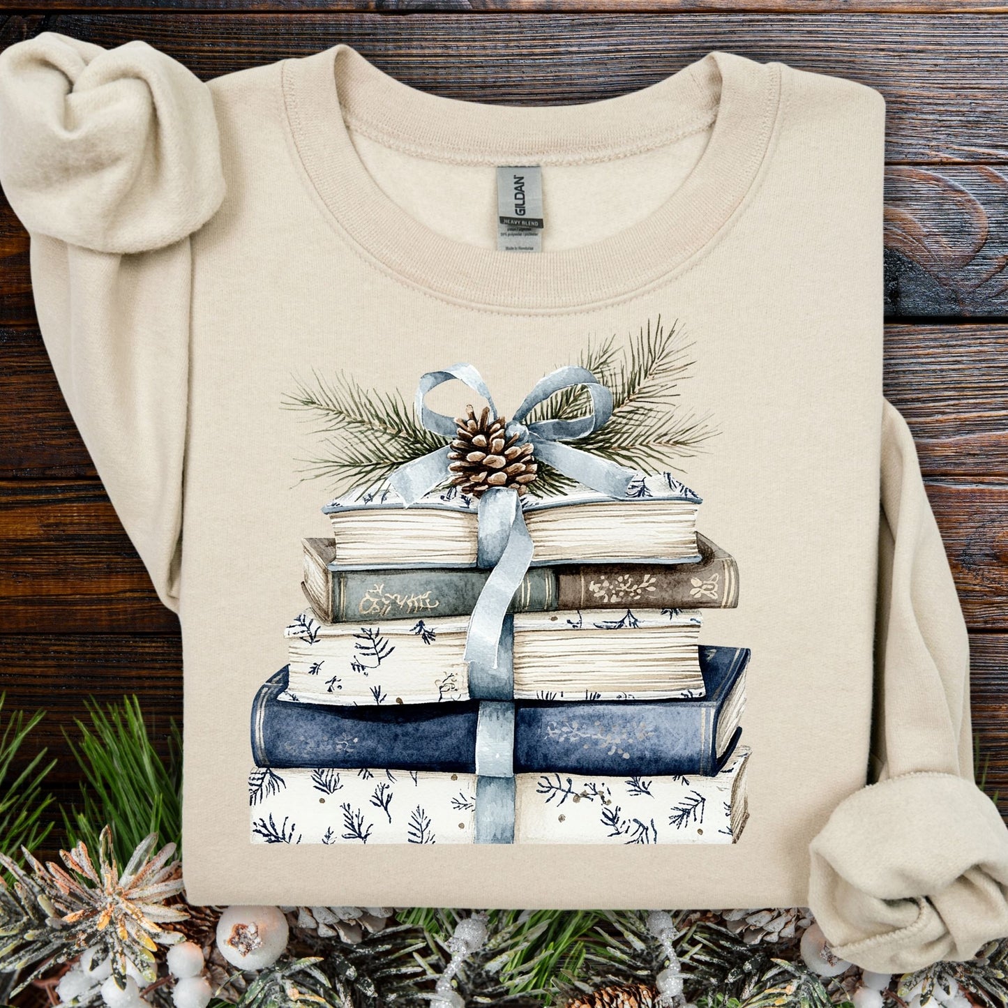 Sand S sweatshirt with blue and white book stack, blue ribbon, evergreen, and pinecone design.