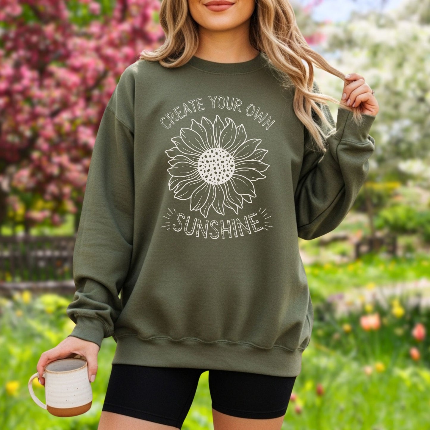 Woman in an olive green crewneck sweatshirt with a white hand-drawn sunflower and "Create Your Own Sunshine" text.