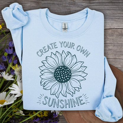 Light blue unisex sweatshirt with "Create Your Own Sunshine" text and a hand-drawn sunflower design.