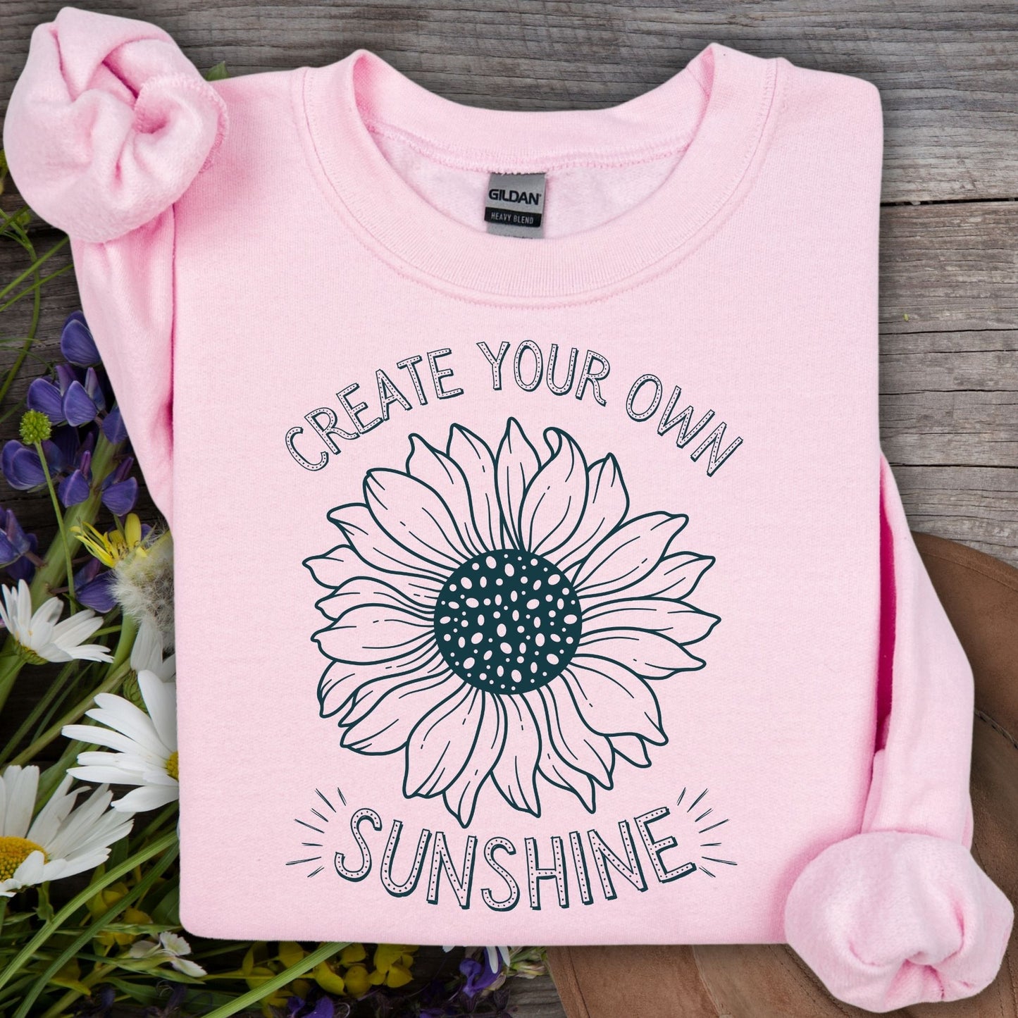 Light pink crewneck sweatshirt with "Create Your Own Sunshine" and a hand-drawn sunflower sketch.