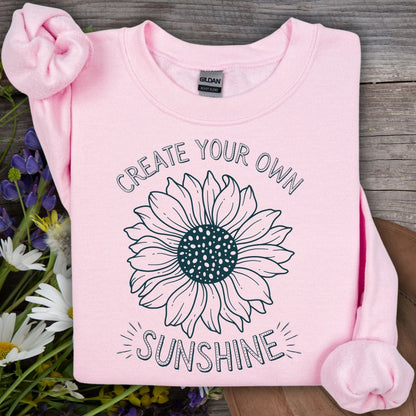 Light pink crewneck sweatshirt with "Create Your Own Sunshine" and a hand-drawn sunflower sketch.