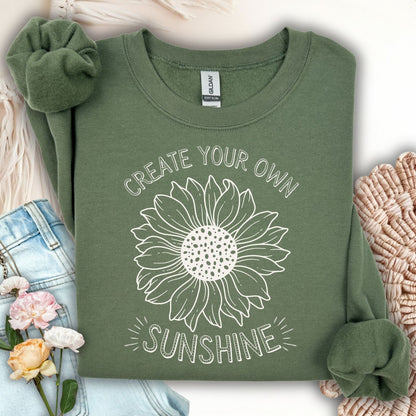 Military green unisex sweatshirt with "Create Your Own Sunshine" text and a hand-drawn sunflower sketch.