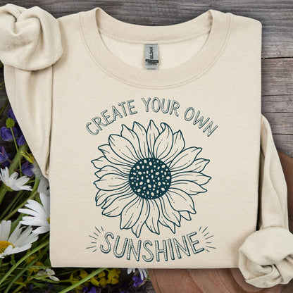 Sand crewneck sweatshirt with a hand-drawn sunflower and "Create Your Own Sunshine" text on a wooden background.