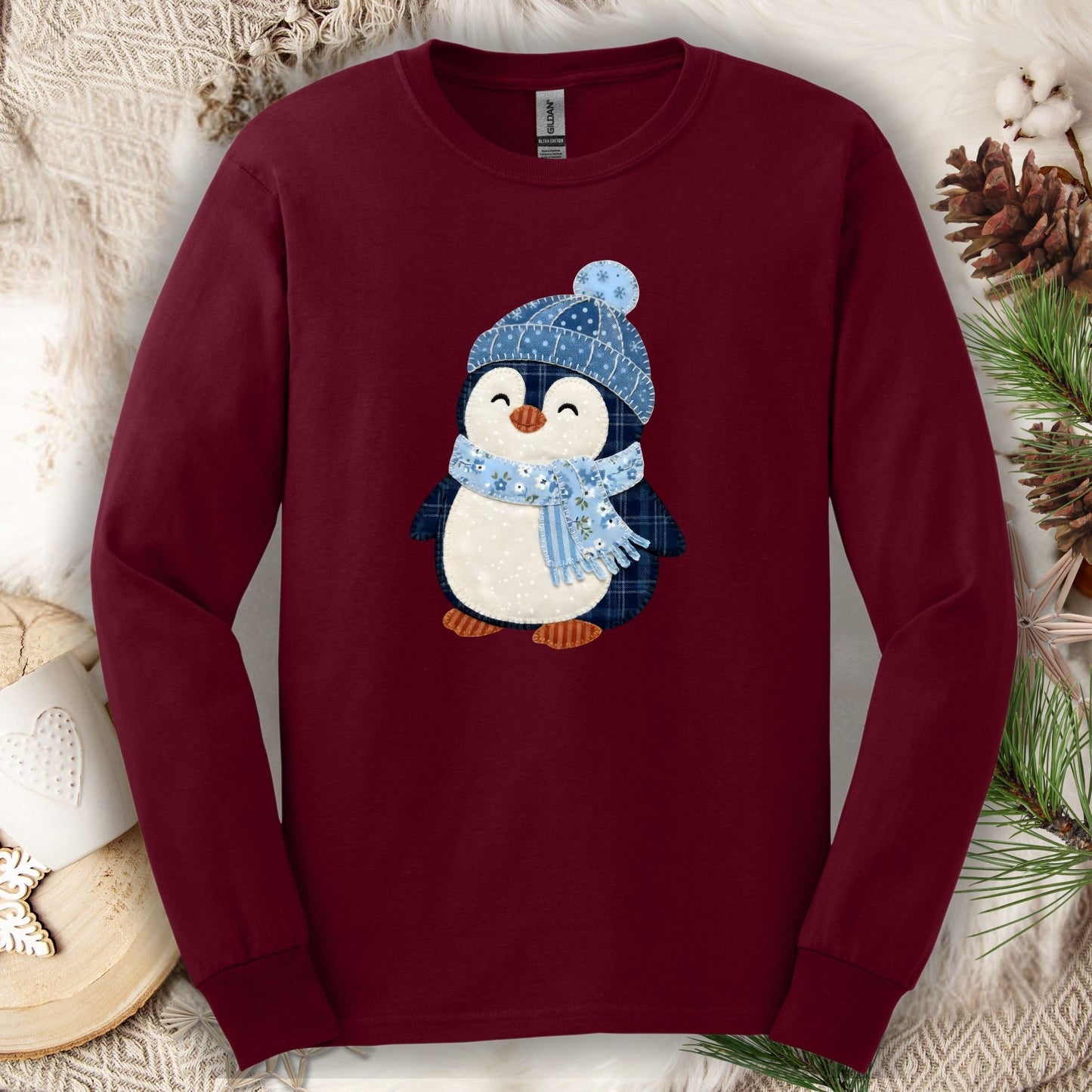 Maroon long sleeve tee with a faux patchwork winter penguin in a blue hat and scarf.