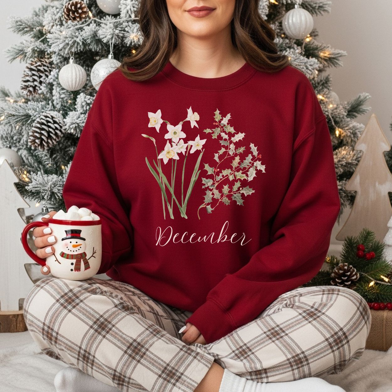 Red crewneck sweatshirt with watercolor narcissus, holly, and "December" text, worn by a person in plaid pajamas.