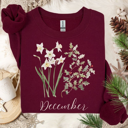 Maroon unisex crewneck sweatshirt with watercolor narcissus, holly, and "December" text, on a cozy winter backdrop.