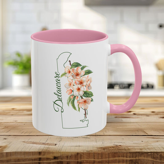 11oz white ceramic mug with pink handle and interior, featuring Delaware state outline and peach blossoms.