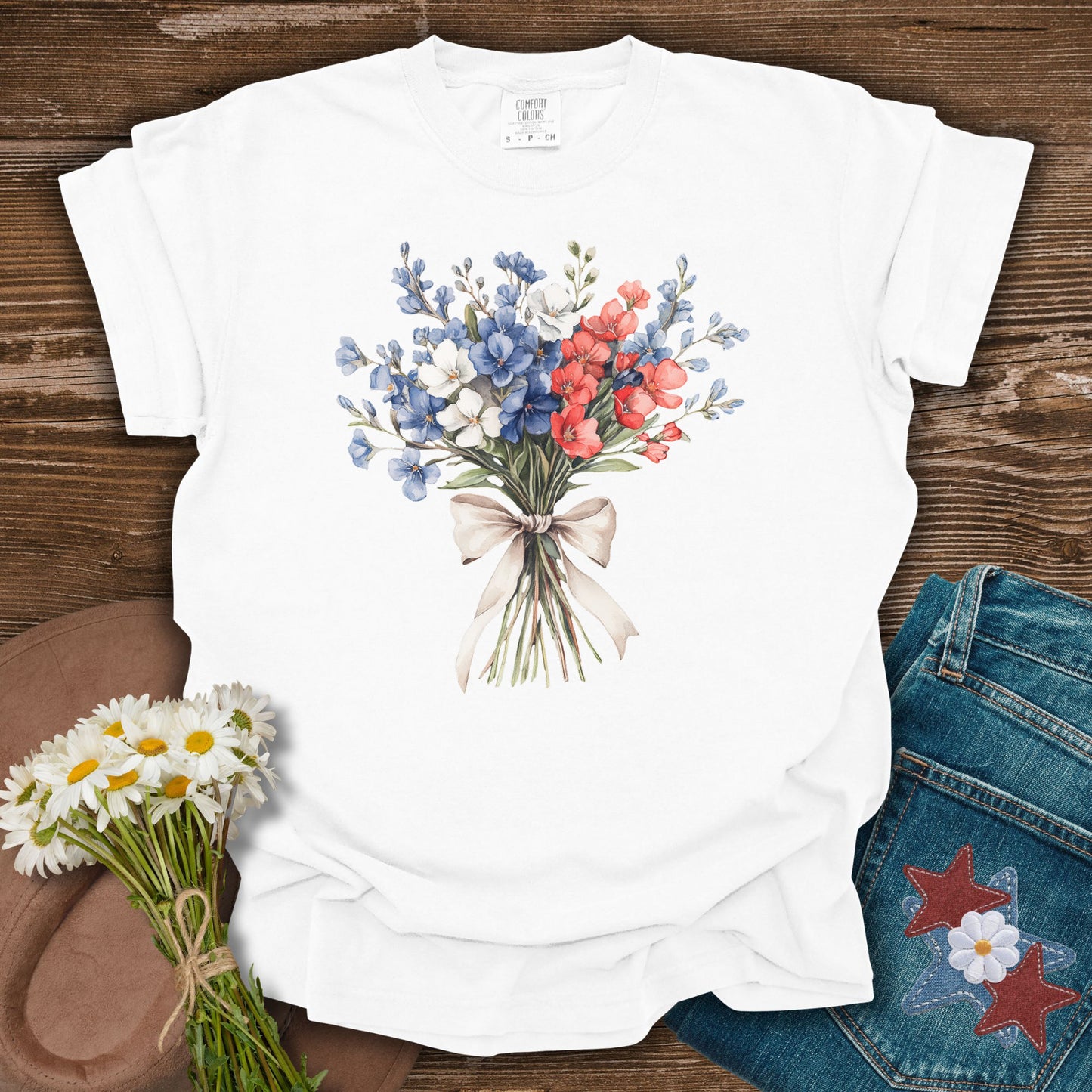 White S t-shirt with a watercolor patriotic bouquet of red, white, and blue flowers tied with a ribbon.