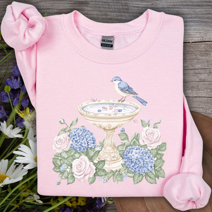 Pink Gildan sweatshirt with a watercolor bird bath, blue bird, roses, and hydrangeas design.
