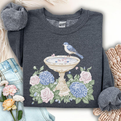 Dark heather sweatshirt with a watercolor bird bath, blue bird, pink roses, and blue hydrangeas.