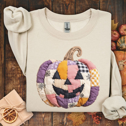 Sand Gildan sweatshirt with a pink and purple patchwork jack-o-lantern design, surrounded by fall decor.