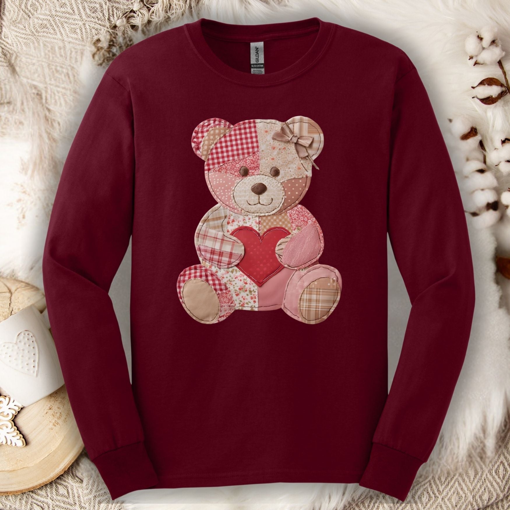 Maroon long-sleeve tee with a faux patchwork teddy bear holding a heart, on a cozy white background.