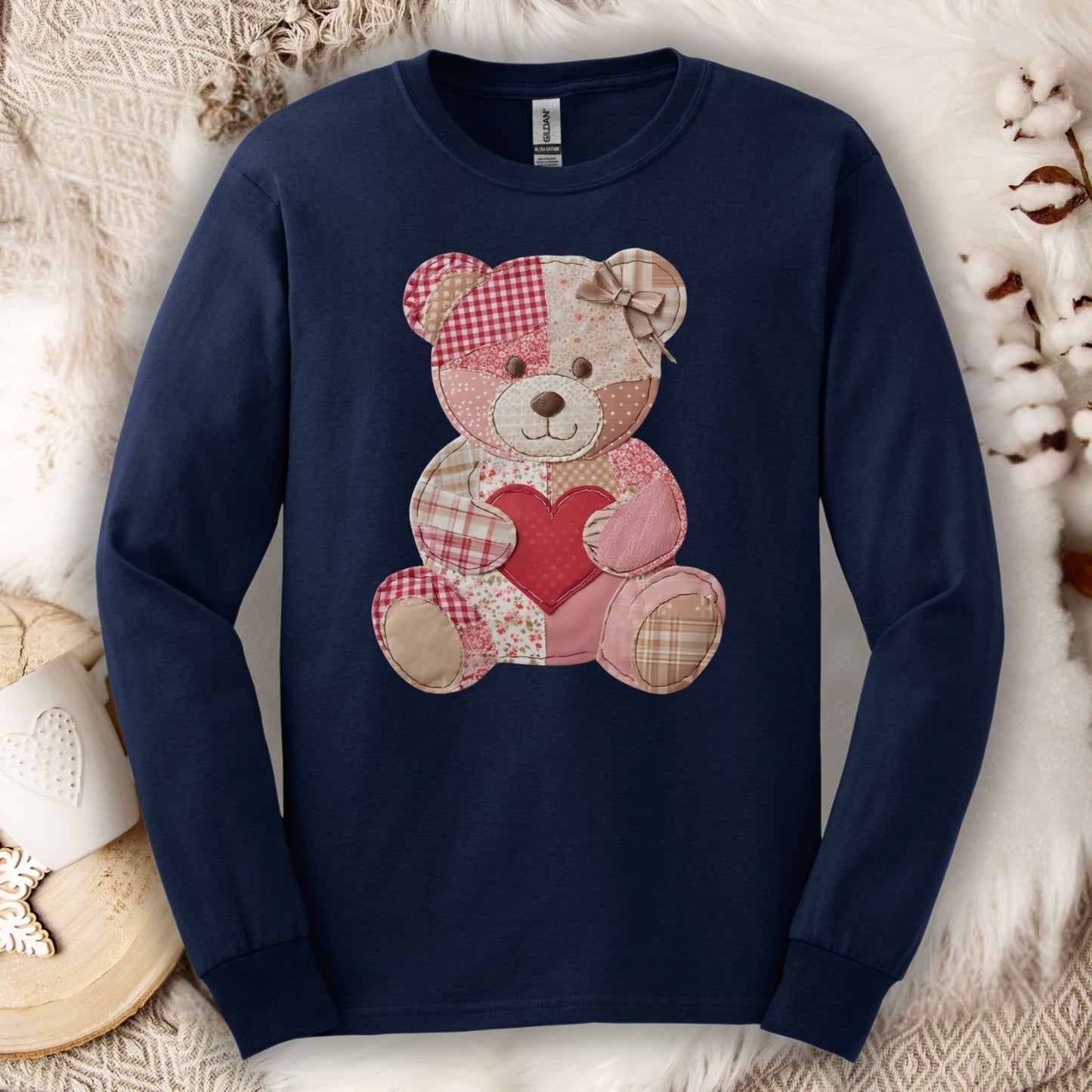 Navy long-sleeve tee with a faux patchwork teddy bear holding a red heart, on a cozy white blanket.