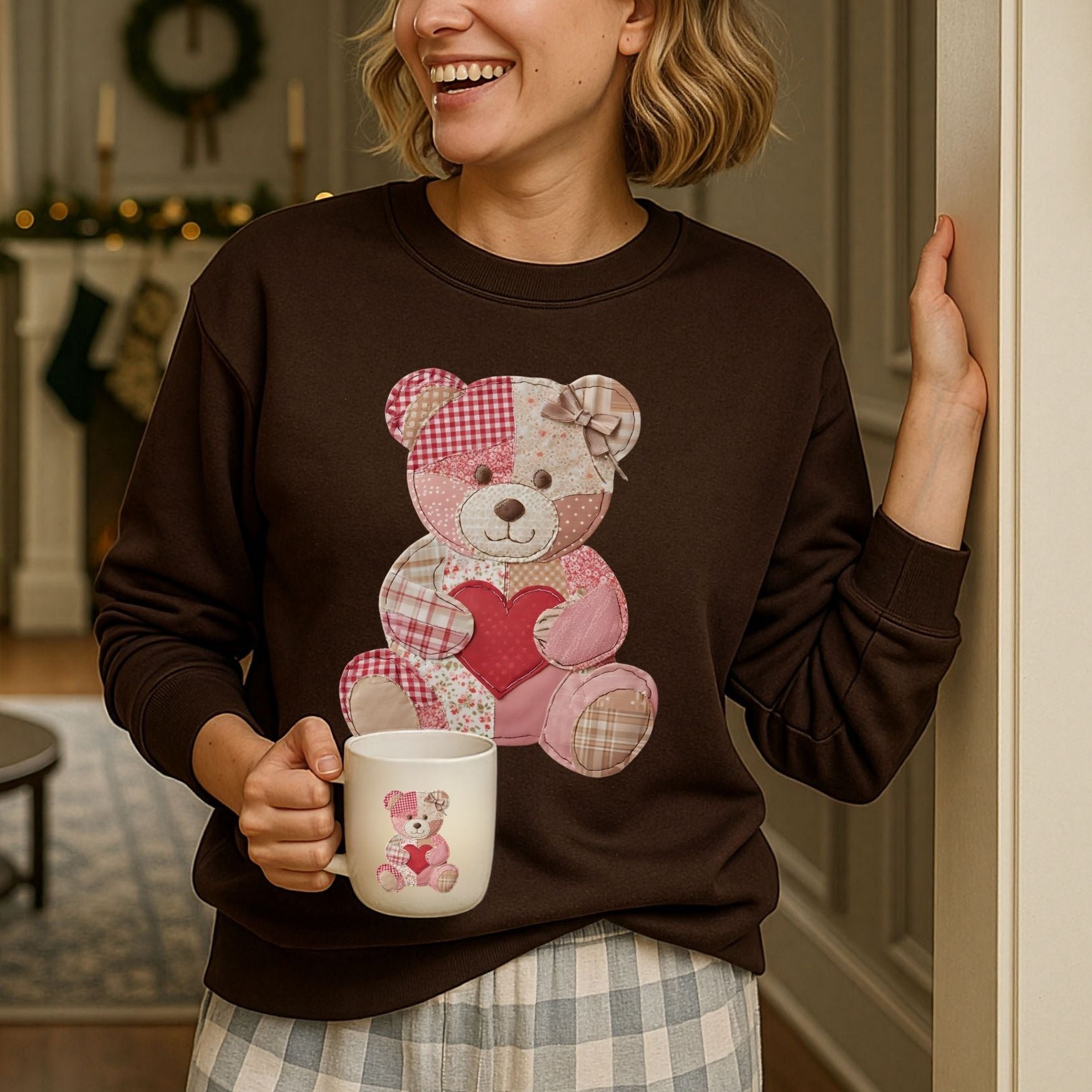 Woman in a brown crewneck sweatshirt with a faux patchwork teddy bear holding a heart, holding a matching mug.