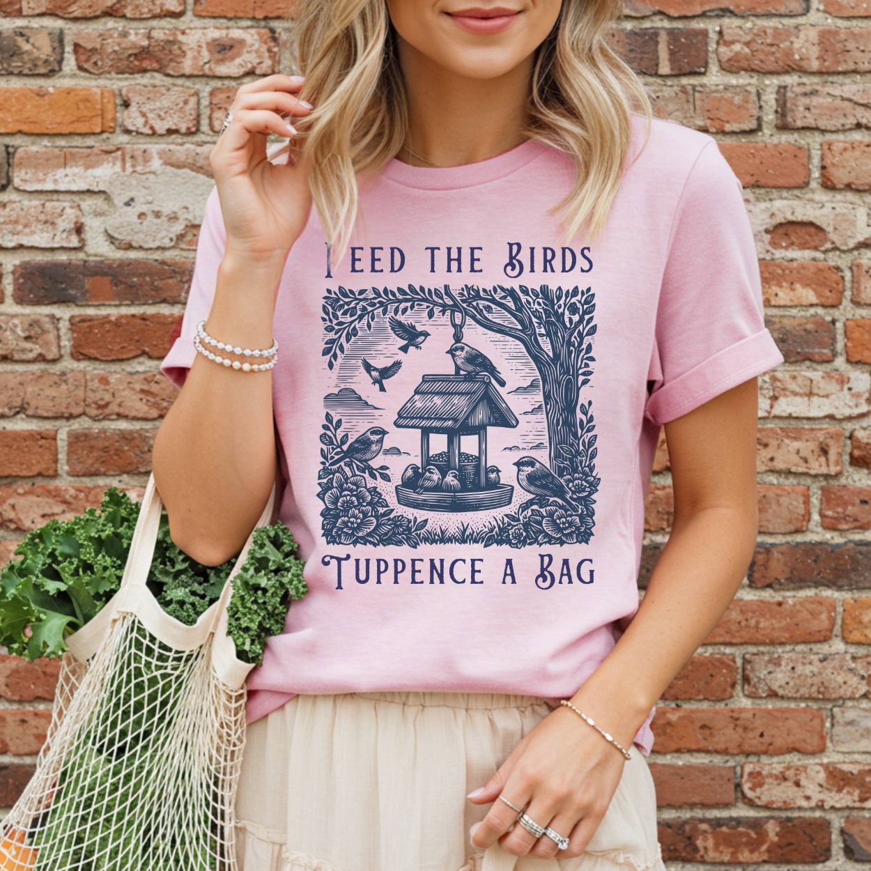 Pink unisex t-shirt with a vintage-style illustration of a bird feeder, finches, sparrows, and "Feed the Birds Tuppence a Bag" text.
